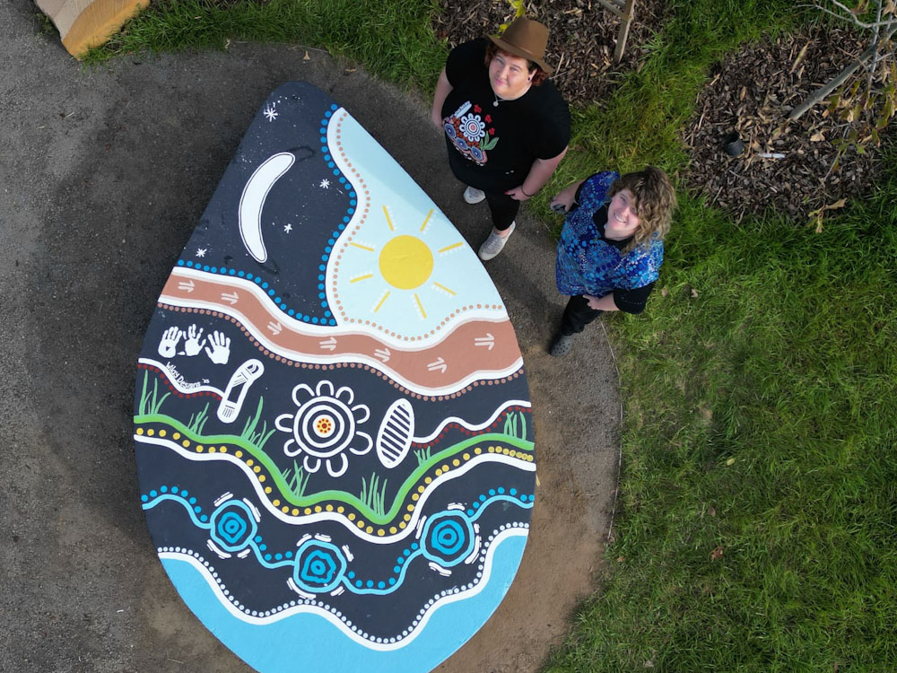 Two Ngunawal sisters and artists, Jazz and Kristal featured with their artwork representing Sky Country. The image is taken from above and looks down on to the painting which covers a concrete plinth in the shape of a teardrop. The plinth is around 2m x 1.5m and around 40cm high. Image features blues, greens, a sun and many Cultural story telling elements on a black background.